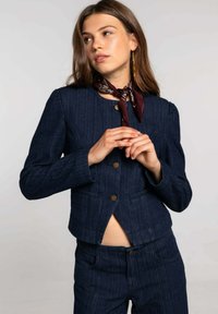 Chaqueta de mezclilla azul oscuro con cuello redondo, cierre de botones y bolsillos frontales. Presenta un corte entallado con un ligero volumen en los hombros.