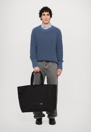 Borsa a tracolla nera con esterno texture, caratterizzata da un dettaglio con logo. Il modello indossa un maglione azzurro a coste e jeans grigi, posando su uno sfondo bianco.