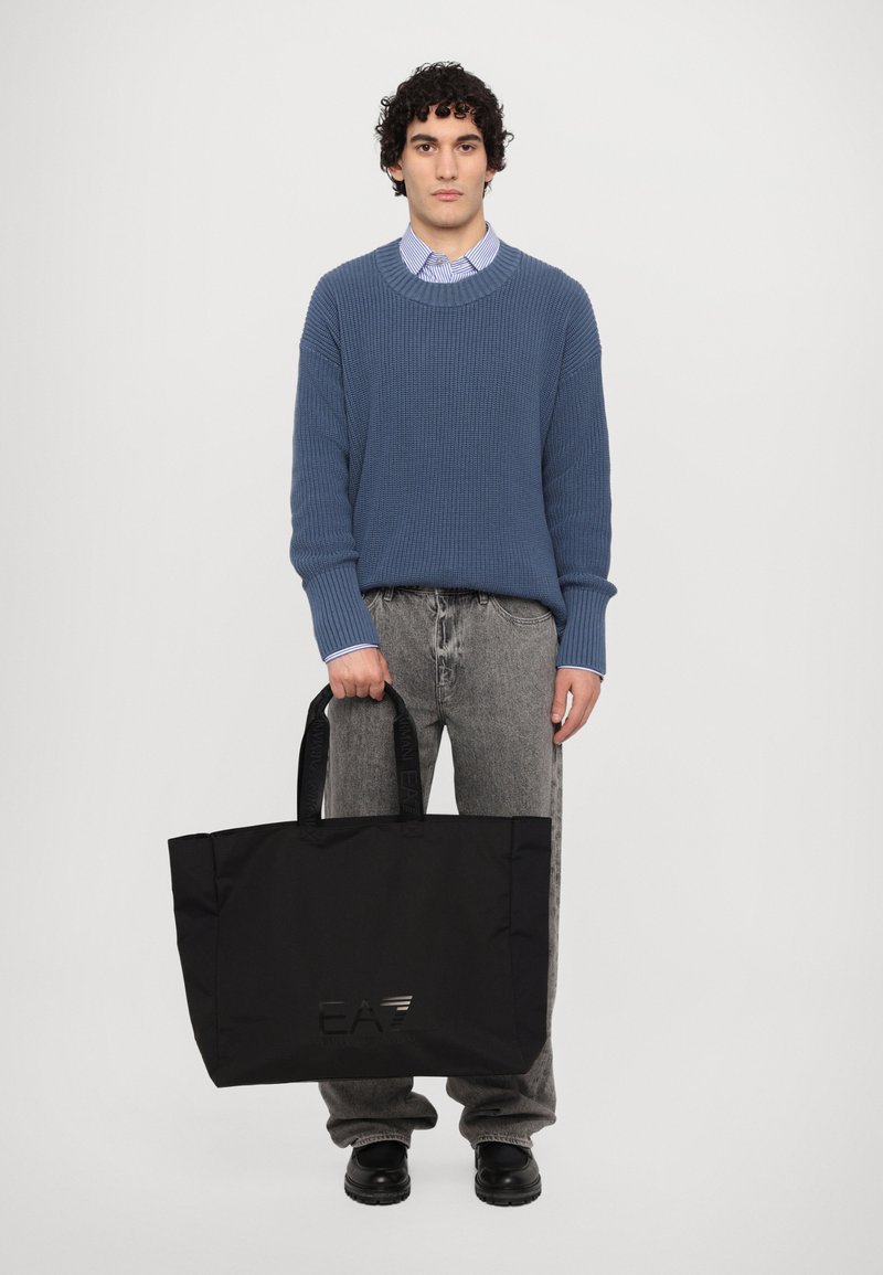 Borsa a tracolla nera con esterno texture, caratterizzata da un dettaglio con logo. Il modello indossa un maglione azzurro a coste e jeans grigi, posando su uno sfondo bianco.