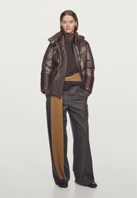 Brązowa pikowana kurtka z wysokim kołnierzem, czarny sweter, szare szerokie spodnie, oraz długi ribbowany brązowy pasek. Błyszcząca struktura kurtki.