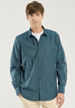 Homme aux cheveux courts et bruns portant une chemise verte sarcelle à manches longues boutonnée sur un t-shirt blanc, pantalon beige, debout les mains dans les poches.