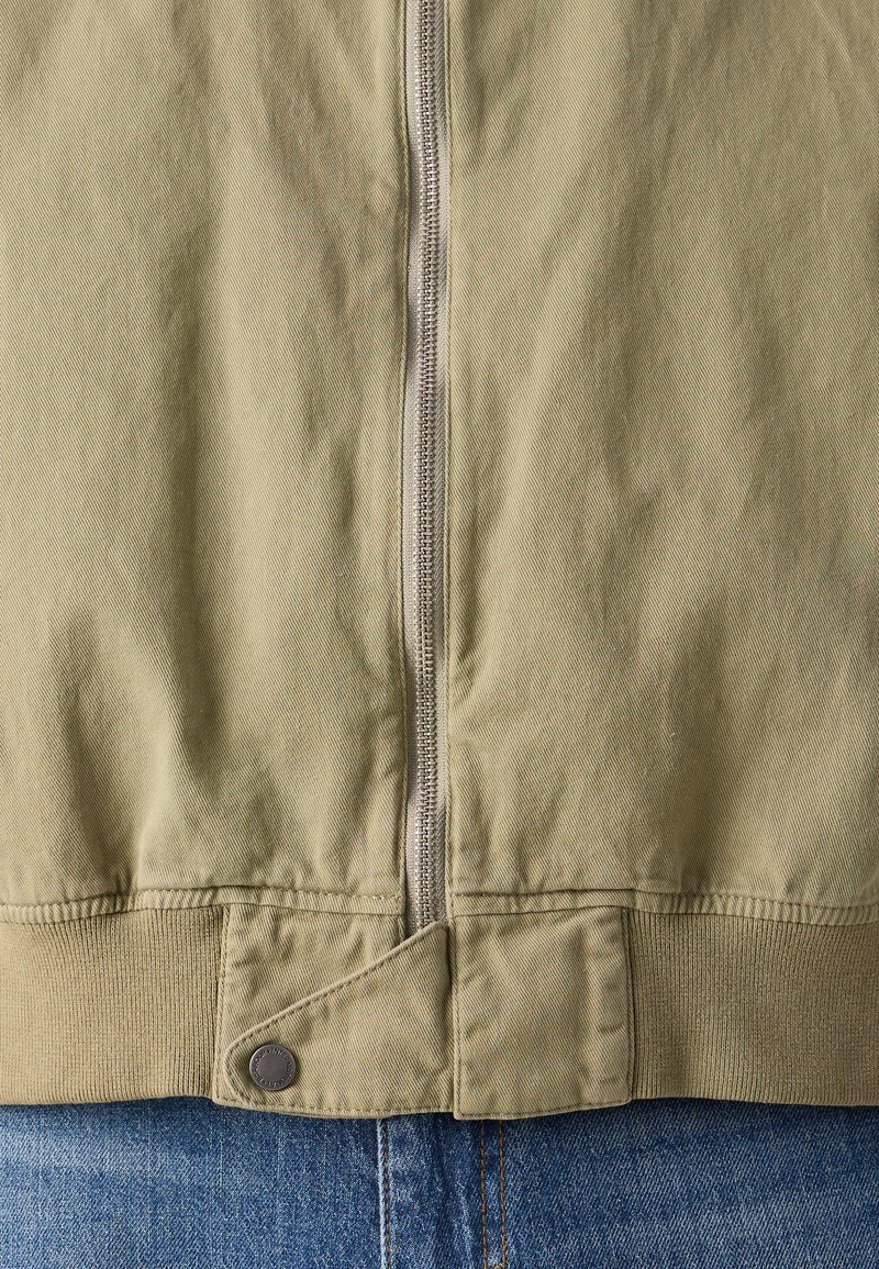 Olivgrüne Bomberjacke mit einem zentralen silbernen Reißverschluss, gerippten Bündchen und Saum sowie einem knopfartigen Akzentdetail am unteren Ende.