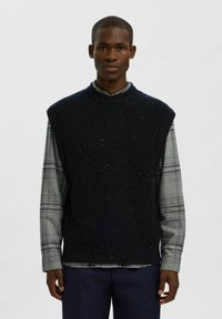 Tamsiai mėlynas be rankovių megztinis su grublėta tekstūra ir purslais, dėvimas ant pilkos langelių marškinės su ilgomis rankovėmis. Paprastas, klasikinis dizainas.