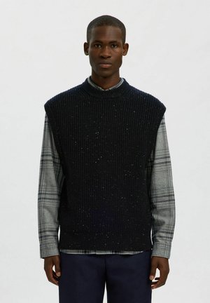 Suéter sin mangas de un azul marino oscuro con textura acanalada y motas, llevado sobre una camisa de cuadros gris con mangas largas. Diseño simple y clásico.
