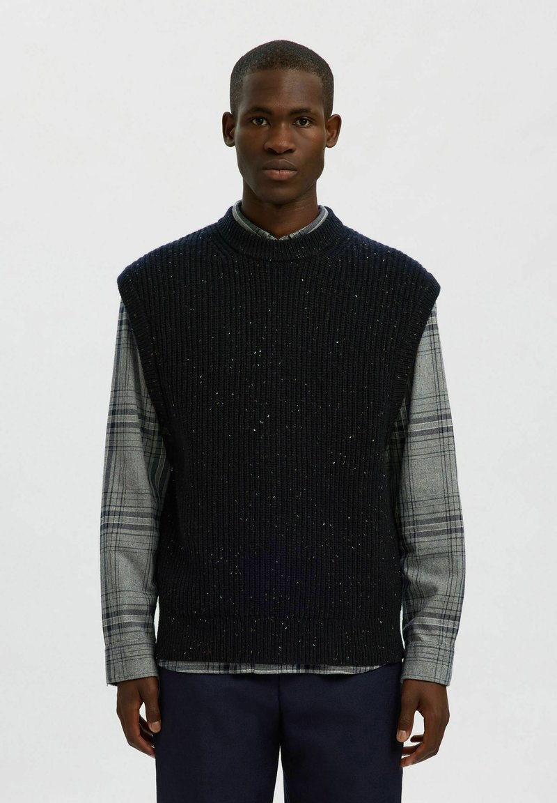 Tamsiai mėlynas be rankovių megztinis su grublėta tekstūra ir purslais, dėvimas ant pilkos langelių marškinės su ilgomis rankovėmis. Paprastas, klasikinis dizainas.