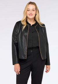 Žena v černé pletené kožené bundě se zlatými řetízky přes černou košili na knoflíky a černé kalhoty, stojící na bílém pozadí.