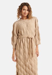 Vestito beige a maglia con motivo a rombi, maniche a palloncino, cintura in vita e spacco laterale per facilitare il movimento.