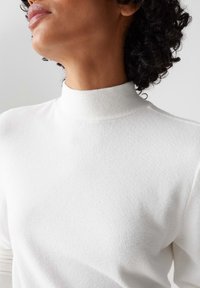 Weißer Pullover mit hohem Kragen aus weichem Stoff, der eine glatte Oberfläche und subtile Nähte am Kragen aufweist.