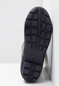 Semelle en caoutchouc noir d'une botte de pluie avec des rainures profondes et le nom de la marque en relief, se tenant debout contre un mur blanc en coin.