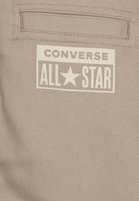 Beżowy materiał płócienny z białą metką, na której widnieje tekst "CONVERSE ALL STAR" oraz wzór gwiazdy. Na brzegach metki widać ściegi.