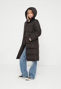 Giacca a vento nera con cappuccio, caratterizzata da una chiusura con zip e tasche laterali. La giacca ha un design trapuntato ed è realizzata in un materiale liscio e resistente.