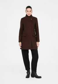 Cappotto di lana marrone con colletto alto, caratterizzato da una chiusura con bottoni nascosti e due tasche a pattina. Abbinato a pantaloni neri e stivaletti.