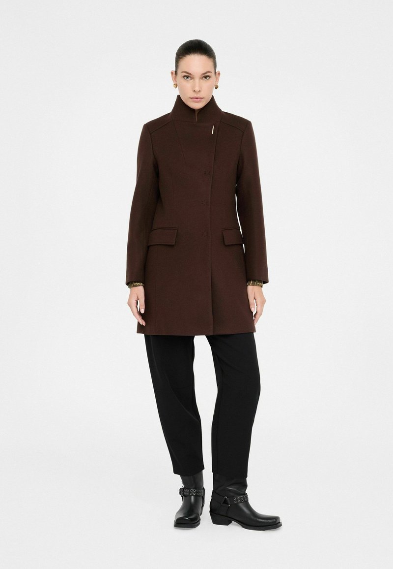 Cappotto di lana marrone con colletto alto, caratterizzato da una chiusura con bottoni nascosti e due tasche a pattina. Abbinato a pantaloni neri e stivaletti.