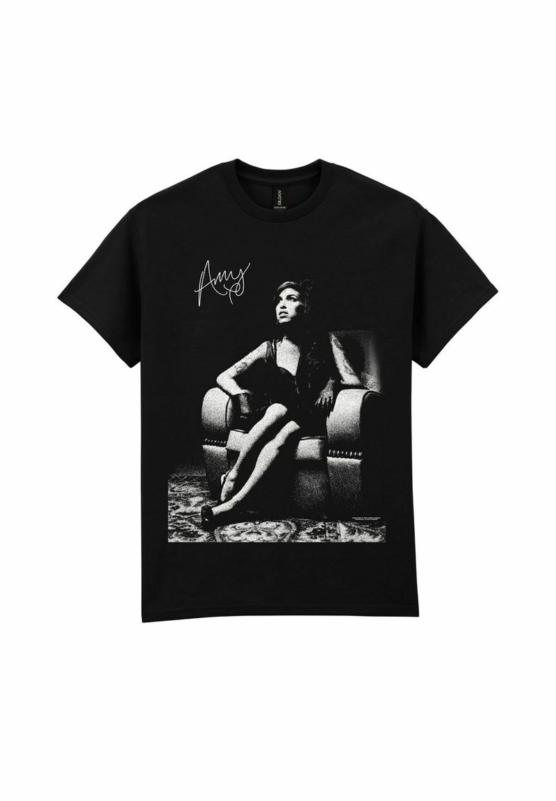 Sort bomulds t-shirt med et monokrom grafisk billede af en siddende kvinde med en signatur ovenover billedet, sat mod en mønstret baggrund.