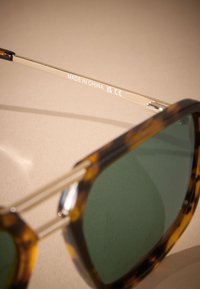 Lunettes de soleil à monture écaille de tortue avec verres verts, branches métalliques, et gravure "FABRIQUÉ EN CHINE" sur un fond clair.