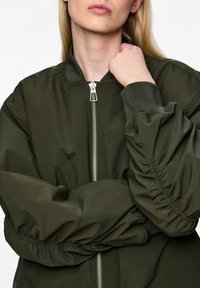 Casaco bomber verde oliva com fecho de zipper à frente, detalhes de mangas franzidas e punhos canelados, exibindo uma textura de tecido leve e suave.