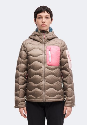 Persoon met kort donker haar draagt een beige gewatteerde jas met capuchon, een roze borstzak, een roze mouwzak en een zwarte broek, kijkt naar voren.