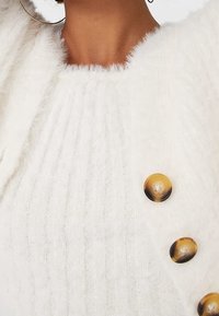 Oberkörper, der einen weißen strukturierten Strickpullover mit großen braunen Verlaufs-Buttons und großen goldenen Creolen trägt.