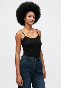 Mulher com um grande cabelo afro, vestindo uma blusa preta sem mangas e jeans azuis, de pé contra um fundo claro simples.