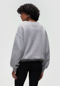 Szary, oversize sweter z opadającymi ramionami i ściągaczami, z małą metką z tyłu. Noszony z teksturowanymi czarnymi legginsami.