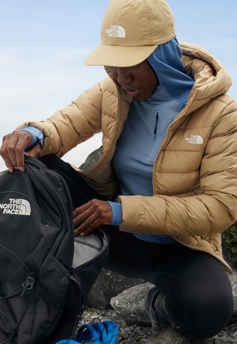 Chaqueta de plumas beige, camiseta de manga larga azul, leggings negros. Mochila negra con logo blanco. Fondo de piedra con cuerda de escalada cercana.
