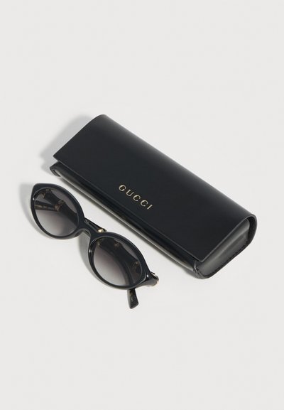 Μαύρα γυαλιά ηλίου σε σχήμα γατίσιου ματιού με φακούς με βαθμιαία απόχρωση, δίπλα σε μαύρη δερμάτινη θήκη Gucci με χρυσά γράμματα, πάνω σε λευκή επιφάνεια.