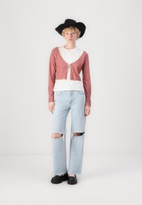 Cardigan scurt cu carouri roșii și albe, cu un nod în față, purtat peste un tricou alb, asortat cu blugi decolorati de un albastru deschis și cizme negre.