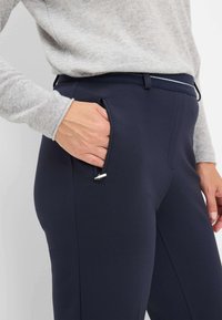 Marineblauwe broek gemaakt van rekbare stof, met een ritszak en een slank ontwerp. Gecombineerd met een lichtgrijze trui, die het detail van de mouwen laat zien.