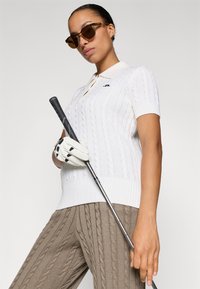 Polo golf a coste bianche con maniche corte, pantaloni beige a coste, guanto da golf bianco e occhiali da sole marroni. Tiene un bastone da golf.