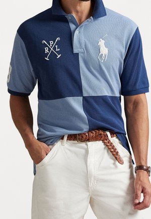 Mand iført en patchwork blå polo med hvid polo spiller emblem, hvide bukser, brun flettet bælte og armbåndsur med læderrem.