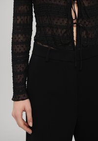 Gros plan sur une personne portant un haut transparent noir à manches longues orné de volants et de liens, associé à un pantalon noir taille haute, avec une position des mains détendue.
