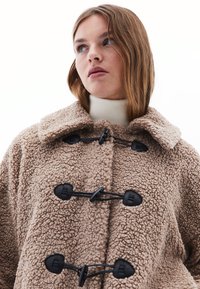 Beige lyhyt takki, joka on valmistettu pörröisestä, teksturoidusta kankaasta. Tässä takissa on kauluspaita ja mustat nyörikiinnitykset edessä.
