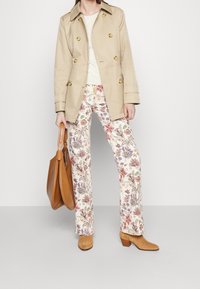 Béžový trenčkot se zlatými knoflíky, krémový top, kalhoty s širokými nohavicemi s květinovým vzorem v pastelových barvách, hnědé boty na podpatku a hnědá taška.