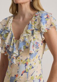 Γυναίκα που φοράει φόρεμα με λουλούδια σε παστέλ αποχρώσεις μπλε, πράσινου και πορτοκαλί, με βολάν στα κοντά μανίκια και στο λαιμό.