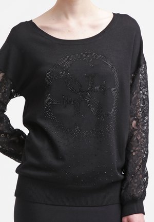 Haut noir à manches longues avec manches en dentelle et un logo circulaire orné de strass à l'avant, porté par une personne.