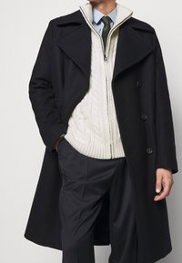 Svart ullrock med breda slag, lager ovanpå en krämfärgad kabelstickad tröja. Mörka byxor fulländar outfiten. Dragkedjan på tröjans krage är synlig.