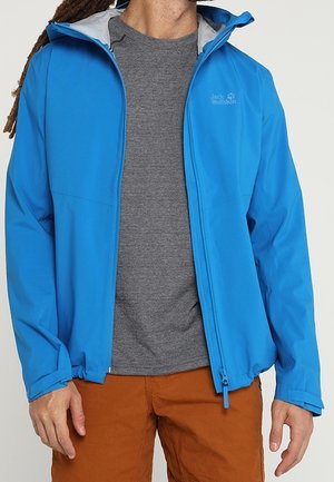 Mann, der eine leuchtend blaue Reißverschlussjacke über einem grauen Hemd und braunen Hosen trägt, steht vor einem schlichten hellen Hintergrund.
