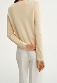 Jasno beżowa bluzka z długim rękawem, wykonana z miękkiego materiału, o minimalistycznym fasonie i okrągłym dekolcie, noszona z białymi spodniami.