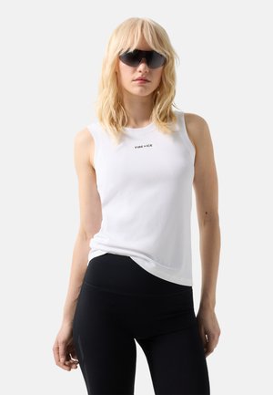 Frau trägt schwarze Sonnenbrille, ein weißes ärmelloses Oberteil mit dem Schriftzug „FIRE + ICE“ und schwarze Leggings, steht vor einem schlichten hellen Hintergrund.