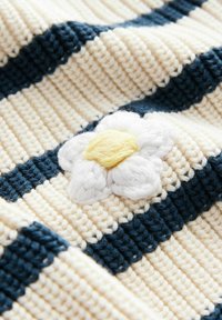 Gestrickte Decke mit cremefarbenen und marineblauen Streifen, verziert mit einer weißen Blumenapplikation und gelbem Zentrum auf der strukturierten Oberfläche.
