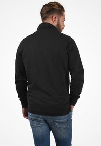 Sudadera negra con cuello alto, mangas largas y puños acanalados, combinada con jeans de mezclilla azul con una textura ligeramente desgastada.