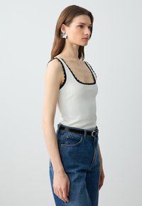 Top in cotone bianco a coste con bordi neri a ondina, abbinato a jeans blu scuro a vita alta e una cintura nera con fibbia argentata.