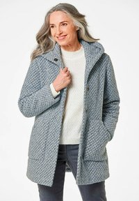 Hellblauer gemusterter Mantel aus strukturiertem Stoff, mit Kapuze, Knopfverschluss und zwei vorderen Taschen, kombiniert mit einem crèmefarbenen Strickpullover.