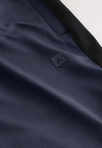 Nahaufnahme eines dunkelblauen Stoffes mit einem kleinen schwarzen quadratischen Logo nahe einer genähten Naht und einem schwarzen Bundabschluss.