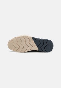 Skoens såle har et sikksakk mønster i beige og marineblått, med kontrasterende teksturer, designet for grep og stabilitet.