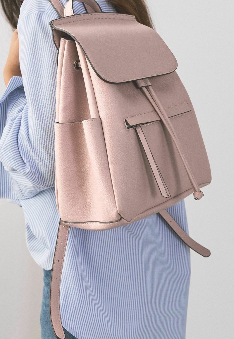 Femme portant un sac à dos en cuir rose clair avec rabat avant et fermeture par cordon, sur une chemise rayée bleu et blanc.