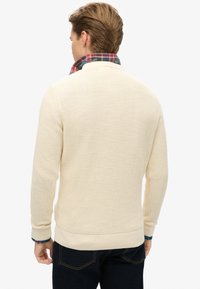 Crème-kleurige gebreide trui met een gestructureerd patroon, voorzien van een hoge kraag en een geruite shirt dat onder de zoom uitspringt. Donkere jeans.