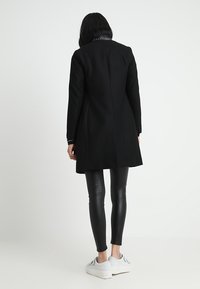 Cappotto nero in lana con una vestibilità sartoriale, caratterizzato da un colletto a contrasto, abbinato a leggings in pelle nera e sneakers bianche.