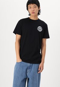 Black cotton t-shirt with a round neckline and short sleeves, featuring a circular graphic logo on the left chest. Worn with blue denim pants.