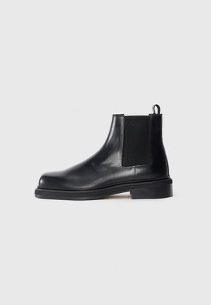 Zwarte leren Chelsea-boot met elastische zijpanelen, ronde neus, aantrekband bij de hiel en een lage gestapelde zool op een effen achtergrond.